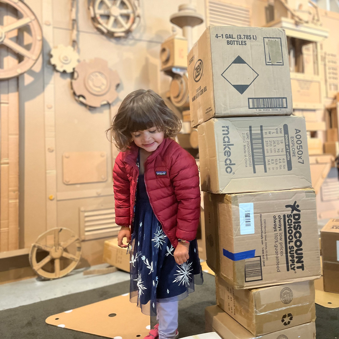 A young child stands next to a stack of cardboard boxes with a cardboard workshop in the background.