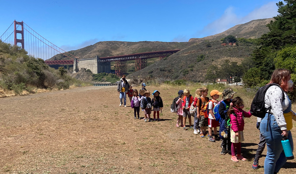 a group of about 15 young campers all dressed in different bright colors walk together with two camp educators. The golden gate bridge is behind them.