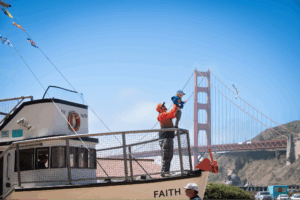A dad holding his son up on the railing of an old fishing boat, with the Golden Gate Bridge in the background