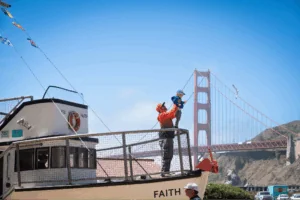 A dad holding his son up on the railing of an old fishing boat, with the Golden Gate Bridge in the background